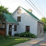Tidy white clapboard house with a green roof and flower beds in Laval